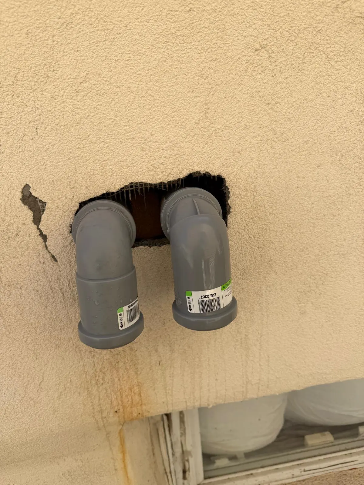 Two drain pipes passing through a large broken hole in a stucco wall