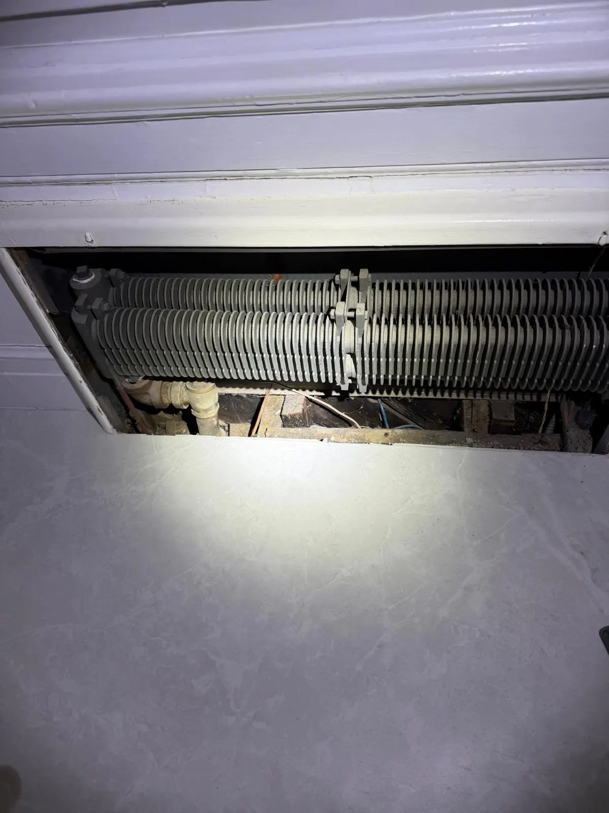 An open cavity beneath a radiator showing exposed floor joists and pipework